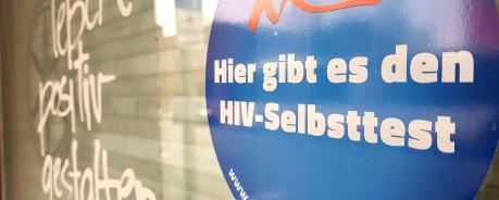 Das Bild zeigt die gläserne Eingangstür der Aidshilfe Osnabrück. Im Vordergrung ist ein Aufkleber zu sehen mit der Aufschrift: Hier gibt es den HIV Selbsttest. Unscharf im Hintergrund der Schriftzug: leben positiv gestalten