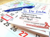 Zu sehen ist eine To Do Liste auf der drei Aufgaben stehen. Durchgestrichen und vermutlich erledigt sind: Aufräumen, Kaffee mit Oma. Rot eingekreist ist die Aufgabe HIV-Test. Die To Do Liste liegt auf einem Kalender. Daneben ist ein Kugelschreiber und im Hintergrund Kondome zu sehen. 