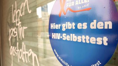 Das Bild zeigt die gläserne Eingangstür der Aidshilfe Osnabrück. Im Vordergrung ist ein Aufkleber zu sehen mit der Aufschrift: Hier gibt es den HIV Selbsttest. Unscharf im Hintergrund der Schriftzug: leben positiv gestalten